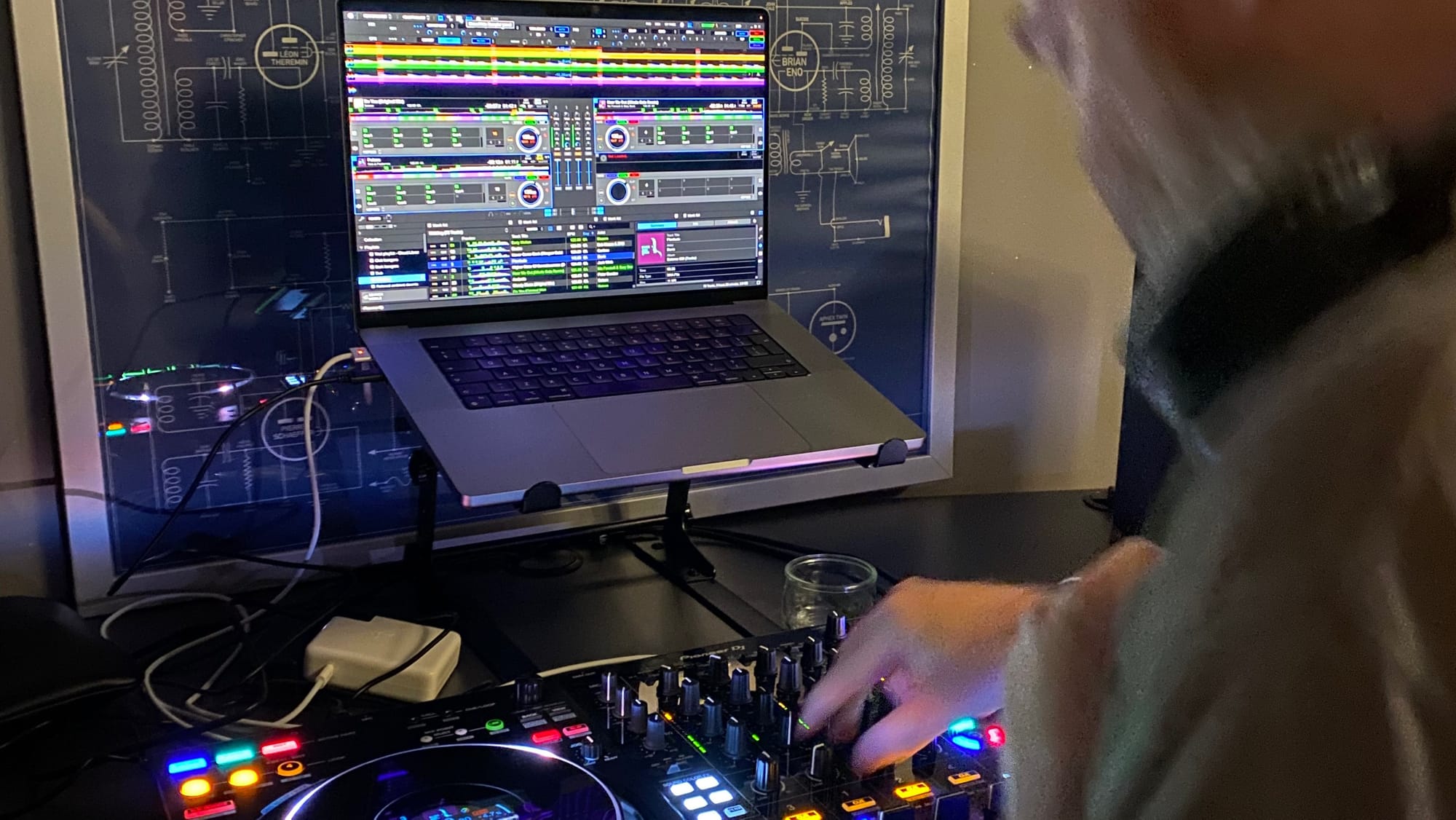 My laptop on a stand, showing the Rekordbox software, with my Pioneer DDJ-FLX10 controller in front, and a blurry me to the right of shot, moving and manipulating some knobs.