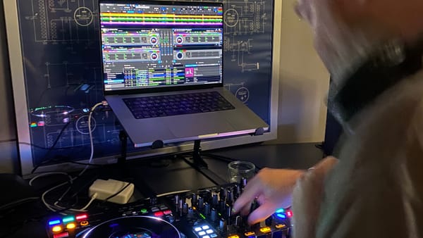 My laptop on a stand, showing the Rekordbox software, with my Pioneer DDJ-FLX10 controller in front, and a blurry me to the right of shot, moving and manipulating some knobs.